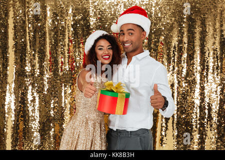 Portrait of smiling young woman in red hats célébrant le Nouvel An tout en maintenant boîte cadeau et geste showing Thumbs up isolated over golden sh Banque D'Images