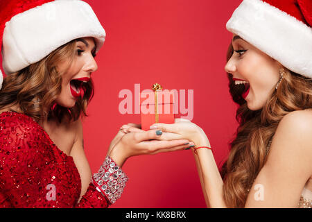 Portrait de deux femmes excitées joyeux noël en tenant un chapeau boîte cadeau et à la recherche à l'autre sur fond rouge isolé Banque D'Images