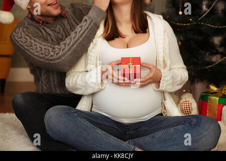 Portrait de femme enceinte avec son mari portant des chapeaux de Noël assis ensemble sur un étage avec un arbre de Noël et présente, l'homme cove Banque D'Images