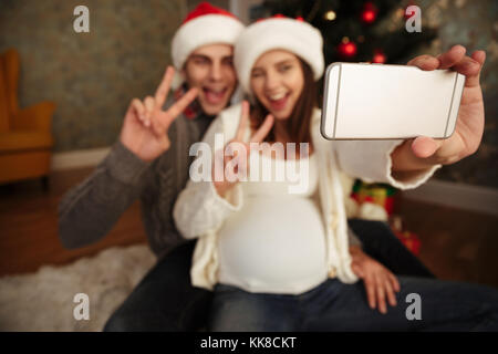 Joyeux sourire femme enceinte avec son mari célébrer Noël en étant assis ensemble sur un étage et prendre une à la maison, couple selfies montrant vic Banque D'Images