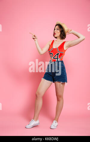 Portrait de jeune femme en rouge étonné et body court jeans tenant son chapeau de paille, en pointant avec le doigt, à côté, isolée sur p Banque D'Images