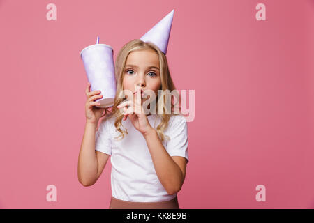 Portrait d'une jolie petite fille dans un anniversaire hat holding cup avec une paille et montrant le silence plus isolés des gestes sur fond rose Banque D'Images