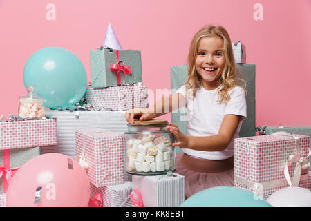 Portrait of a happy little girl dans un chapeau d'anniversaire célébrant tout en étant assis sur une pile de boîtes actuelle avec un pot de guimauve rose plus isolés b Banque D'Images