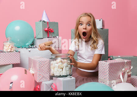 Portrait d'une petite fille choquée dans un chapeau d'anniversaire célébrant tout en étant assis sur une pile de boîtes actuelle avec un pot de guimauve rose plus isolés Banque D'Images