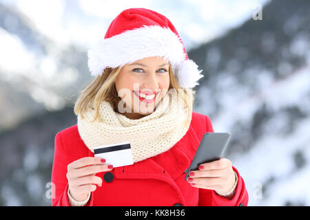 Happy girl sur le port de noël santa claus hat holding credit card et smart phone avec la montagne enneigée en arrière-plan Banque D'Images