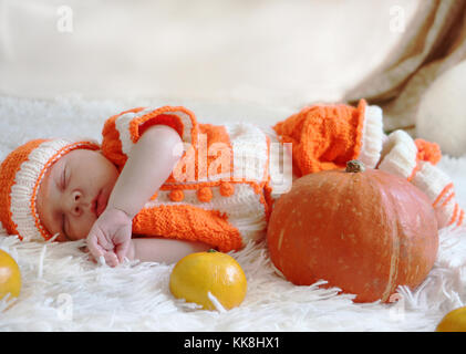 Nouveau-né en costume orange tricoté dormir sur couverture blanche à la citrouille et des oranges en face de lui. Thanksgiving Halloween ou concept. Banque D'Images