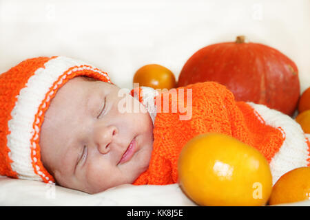 Cute smiling couchage bébé nouveau-né vêtu d'un costume orange tricoté à la citrouille et des oranges. La récolte ou de l'automne halloween concept. Smi Banque D'Images