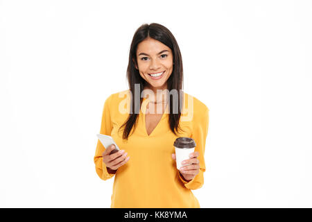 Close-up portrait of happy young brunete femme en chemise jaune holding tasse de café et téléphone mobile, looking at camera, isolated over white backgrou Banque D'Images