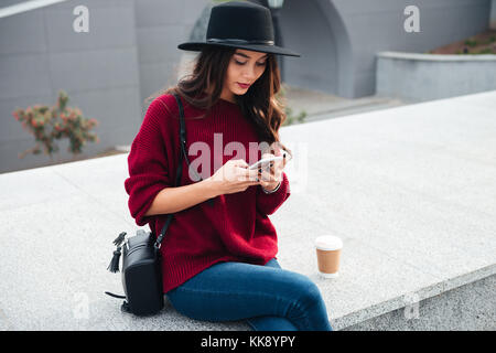 Portrait of a beautiful young asian girl wearing hat chandail et à l'aide de téléphone mobile tout en étant assis sur une rue de la ville Banque D'Images