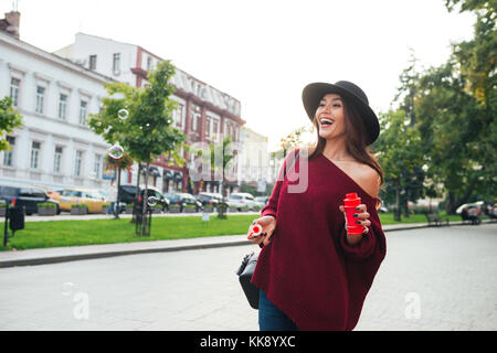 Portrait of a smiling happy asian woman in hat soufflant des bulles de savon en se tenant debout à l'extérieur, sur une rue de la ville Banque D'Images