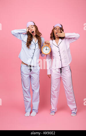 Photo de deux amis en pyjama béant femmes isolées sur fond rose holding réveil. Banque D'Images