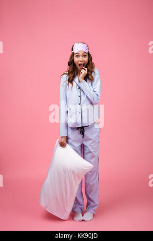 Portrait d'une jeune fille choquée en pyjama holding pillow debout et crier plus isolé sur fond rose Banque D'Images