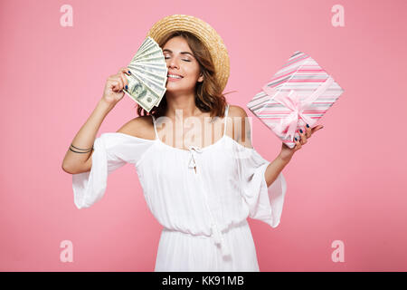 Photo d'une belle jeune femme au chapeau de paille, avec les yeux fermés, holding dollar bills and gift box, isolé sur fond rose Banque D'Images