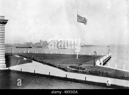 Une photographie montrant une partie de la pelouse qui entoure la station d'inspection de l'immigration sur Ellis Island, le bâtiment de style Renaissance française est construite avec des briques rouges et de calcaire de caisse, le bâtiment a ouvert ses portes le 17 décembre 1900, il a été fermé en 1954, après le traitement de plus de 12 millions d'immigrants qui entrent aux États-Unis, un indicateur est utilisé à l'extérieur du bâtiment, une grande promenade est séparée de l'herbe par des clôtures basses et des arbustes, la ville de New York peut être vu dans le contexte de l'autre côté de la baie de New York, 1907. À partir de la Bibliothèque publique de New York. Banque D'Images