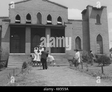 Une photographie d'un groupe d'huissiers et un photographe de l'église, les filles du groupe porter des robes blanches identiques alors que le seul garçon porte un costume et une cravate, ils sont entre des photographies comme une femme en blanc travaille pour ajuster le positionnement du groupe, le photographe est mis en place directement en face du groupe qui posent sur les étapes menant à l'entrée de l'église, deux adultes regardez sur la pelouse de l'église, Durham, North Carolina, 1940. À partir de la Bibliothèque publique de New York. Banque D'Images