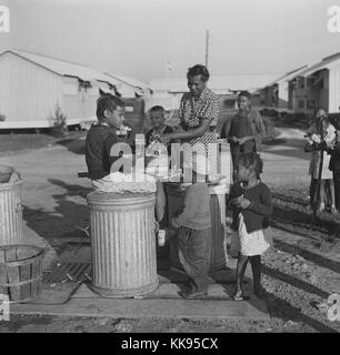 Photographie en noir et blanc d'une femme afro-américaine, avec des enfants, lave-verts par près de la pompe d'abris en métal à Okeechobee, Camp de travail migrateurs Belle Glade, en Floride, Février, 1941. À partir de la Bibliothèque publique de New York. Banque D'Images