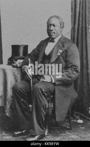 Un portrait photographique d'un homme non identifié, il est vêtu d'un costume de couleur sombre, chemise blanche et noeud papillon, il est assis avec son bras reposant sur une table sur laquelle il a placé son chapeau, il tient une canne, il est positionné à un angle de 45 degrés à l'appareil photo, 1900. À partir de la Bibliothèque publique de New York. Banque D'Images