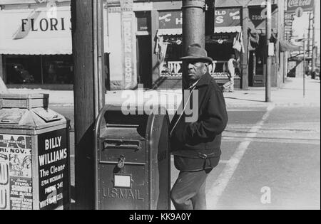 Une photographie d'un homme debout par un United States Postal Service drop box sur coin de rue, l'homme porte une veste de couleur sombre, un pantalon et un chapeau, une corbeille est situé à côté de la boîte de dépôt et contient des publicités pour des entreprises locales et des événements, les magasins peuvent être vus de l'autre côté de la rue directement derrière l'homme, Newport News, Virginie, 1940. À partir de la Bibliothèque publique de New York. Banque D'Images