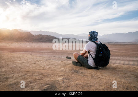Le port du foulard voyageur arabe avec un sac à dos, s'assoit et regarde les montagnes et le désert du Sinaï, en Égypte, au coucher du soleil Banque D'Images