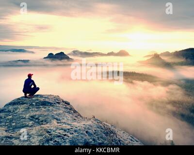 Moment de solitude sur le sommet rocheux exposés. man in black profitez d'une vue merveilleuse. s'asseoir sur le randonneur pic de roche et de regarder dans la brume et le brouillard coloré Banque D'Images