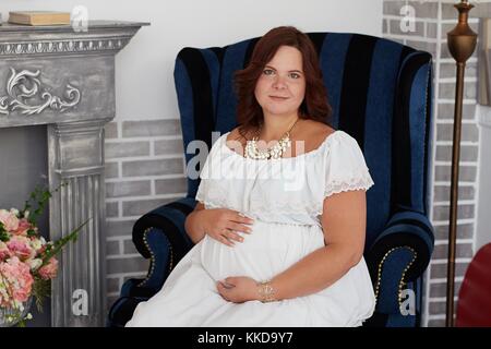Portrait de femme enceinte avec adorable sourire heureux dans la pure robe blanche et des bijoux embrasse son ventre, assis dans un fauteuil près de cheminée vintage Banque D'Images