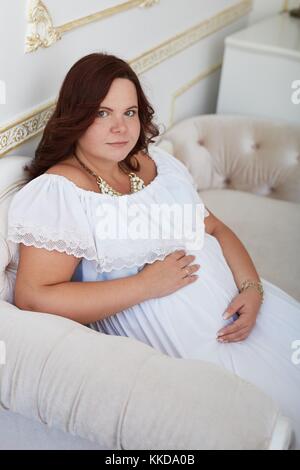 Belle femme enceinte pacifique avec sourire heureux dans la pure robe blanche et des bijoux mis sa main sur ventre assis sur un canapé blanc dans la décoration de luxe int Banque D'Images