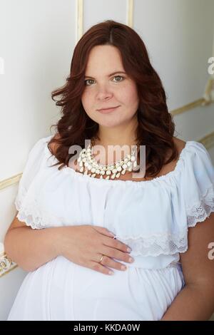 Belle femme enceinte avec sourire heureux et paisible visage habillé en robe blanche et pure bijoux mit sa main sur le ventre Banque D'Images