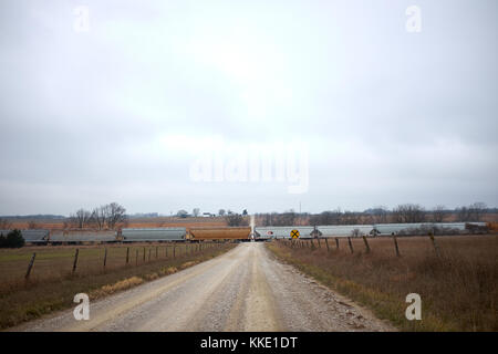 Long train de fret traversant une route de terre rurales éloignées qui traverse les terres agricoles en vue d'un recul tout droit sous les nuages gris Banque D'Images