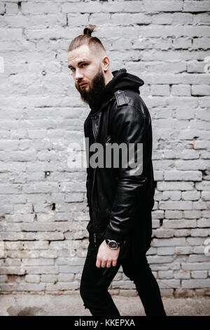 Jeune homme barbu moderne en noir vêtements de style posant contre un mur de briques. Banque D'Images