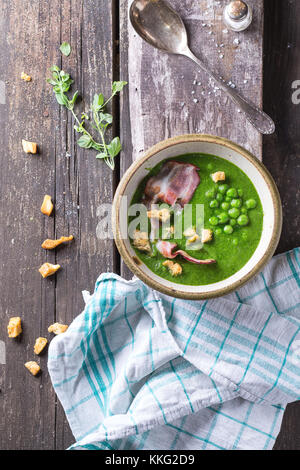 Soupe crème de petits pois dans un bol en céramique avec lard grillé et croûtons. plus vieille table en bois avec des serviettes de cuisine, vintage cuillère, grenier à sel et d'herbes Banque D'Images