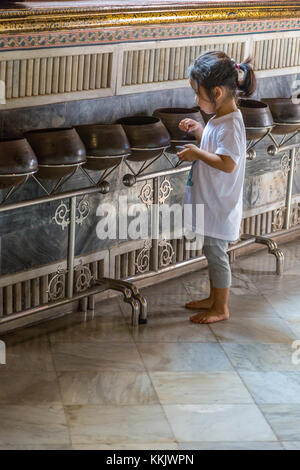 Bangkok, Thaïlande. Wat Pho temple du Bouddha couché. Petite fille en plaçant des pièces de monnaie dans des pots tout en cherchant des bénédictions ou la bonne fortune. Banque D'Images