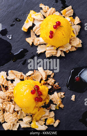 La crème glacée, sorbet mangue servi sur les miettes de gaufrettes avec baies de groseille rouge et de cubes de glace sur noir ardoise. vue d'en haut. Banque D'Images