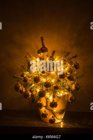 Petit arbre de Noël avec des lumières chaudes et babioles de luxe, dans un pot en plastique blanc, avec le fond sombre. Banque D'Images