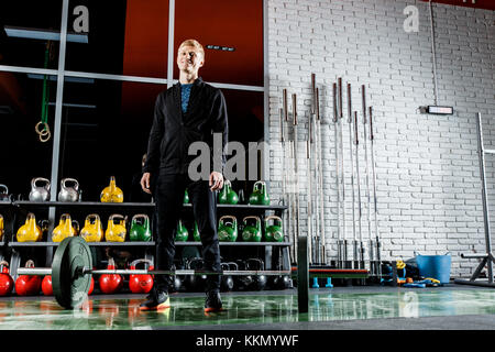 Le mec soulève le bar dans la salle de sport sur l'arrière-plan d'une fenêtre et un mur de brique gris Banque D'Images