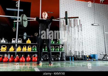 Le mec soulève le bar dans la salle de sport sur l'arrière-plan d'une fenêtre et un mur de brique gris Banque D'Images