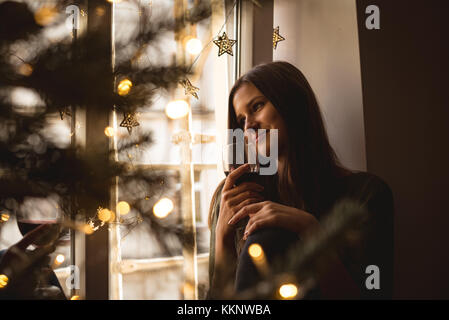 Femme regardant par la fenêtre tout en ayant un verre de vin pendant noël Banque D'Images