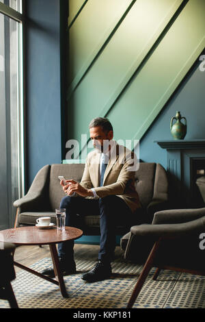 Voir à l'homme d'âge moyen de boire du café et à l'aide de téléphone mobile dans le lobby Banque D'Images