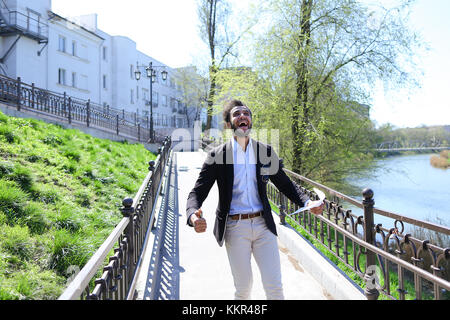 Belle personne parle par téléphone et la marche sur le pont. l'homme met les mains en l'air joyeusement avec papiers. Guy a des cheveux noirs courts, barbe, lunettes de soleil sur une poche Banque D'Images