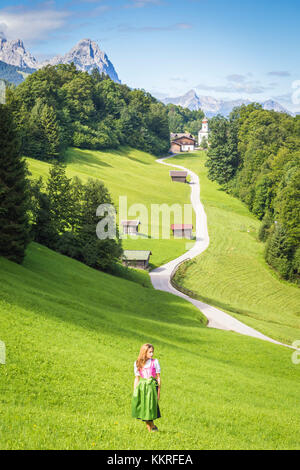 Une fille en robe typique du Bayern marchant devant le village de Wamberg, avec le mont Zugspitze et Waxenstein en arrière-plan. Garmisch-Partenkirchen, Bayern, Allemagne. Banque D'Images