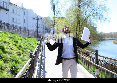 Belle personne parle par téléphone et la marche sur le pont. l'homme met les mains en l'air joyeusement avec papiers. Guy a des cheveux noirs courts, barbe, lunettes de soleil sur une poche Banque D'Images