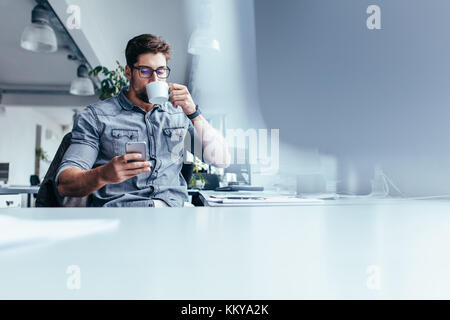 Designer hommes de boire du café tout en lisant un message sur téléphone intelligent. Jeune homme à la tasse de café et à la recherche au téléphone mobile. Banque D'Images