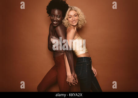 Portrait de deux jeunes femmes se tenant debout dos à dos et à la recherche à l'appareil photo sur fond brun. Femelle multi ethnic des modèles posant en studio et souriant. Banque D'Images