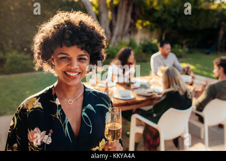 Smiling african woman standing en plein air avec des gens assis en arrière-plan ayant la nourriture à partie. Femme avec un verre au partie en plein air. Banque D'Images