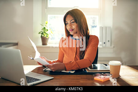 Souriante jeune femme entrepreneur africain assis à une table dans son bureau à domicile la lecture de documents et de travailler sur un ordinateur portable Banque D'Images
