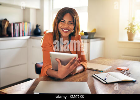 Souriante jeune femme africaine en ligne de travail avec une tablette numérique tout en restant assis à sa table de cuisine à domicile Banque D'Images