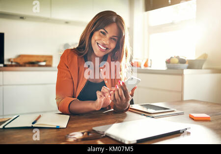 Souriante jeune femme entrepreneur africain travaillant à table dans sa cuisine à la maison d'envoyer des messages texte sur son portable Banque D'Images