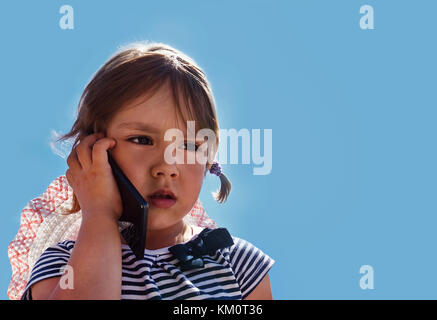 Petite fille avec un regard triste et malheureux yeux parler au téléphone avec ma mère sur le fond de ciel bleu à l'extérieur dans le parc. Banque D'Images