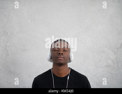 Portrait d'un homme à la peau sombre, les yeux fermés, écouteurs, a l'écoute de musique préférée. adolescent bénéficie d'une piste audio isolée sur mur blanc Banque D'Images