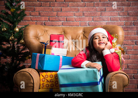 Little smiling indian girl with christmas gifts lors du port de santa claus dress et assis sur fauteuil ou canapé et l'arbre de Noël en arrière-plan Banque D'Images