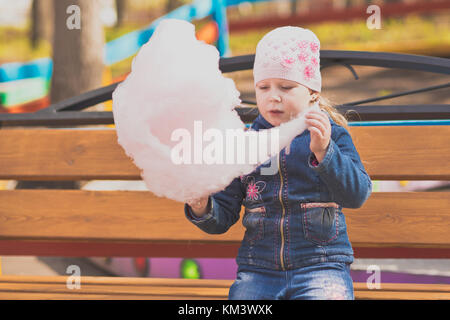 Petite fille sur un banc de parc eating cotton candy. portrait d'une jeune fille dans le parc sur une marche. Banque D'Images